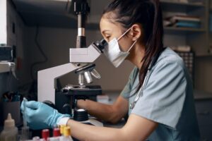 Asian woman with medical mask looks into microscope at table in hospital